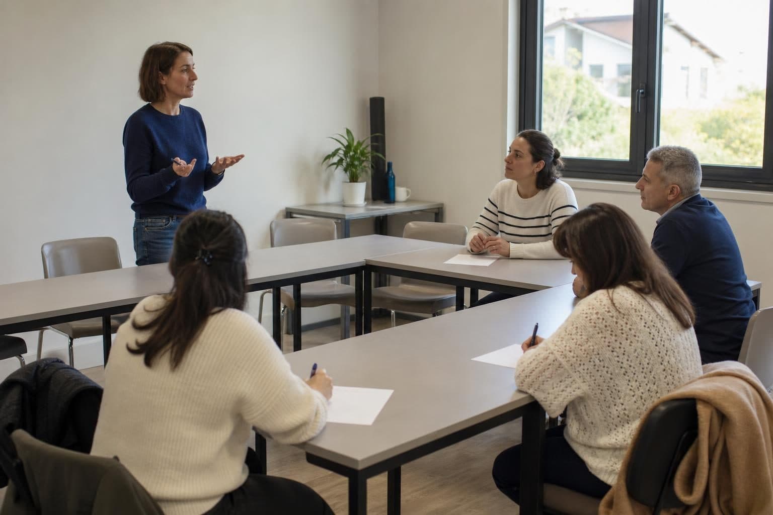 Une formatrice se tient debout devant un petit groupe assis autour de tables en U dans une salle de formation lumineuse.