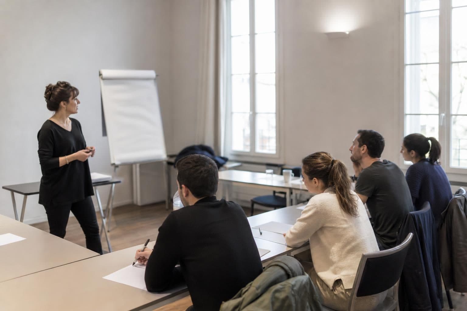 Une formatrice debout anime une petite session de formation devant plusieurs adultes assis autour de tables dans une …